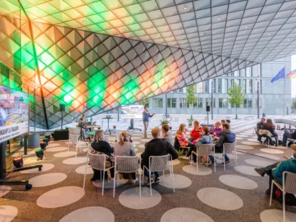 Futurium Vorplatz. Menschen sitzen verteilt an kleinen Tischen. Die Decke ist mit bunten Farben beleuchtet und lenks an der Wand steht ein großer Fernseher.