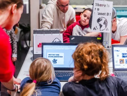 Zwei Kinder designen ein Plakat am Laptop. Darauf steht "Rettet die Schildkröten".