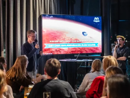 Zwei männliche Moderatoren stehen vor einem großen Fernseher. Vorne im Bild ist ein Publikum zu sehen. Die Aufnahme stammt von einem Kneipenquiz.