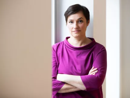 Nicole Deitelhoff steht mit verschränkten Armen vor einem hellen Hintergrund. Sie trägt ein fliederfarbenes Oberteil.