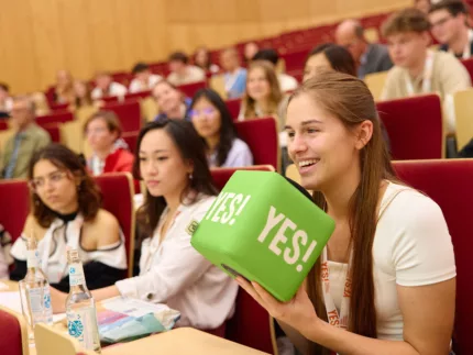 Auf dem Bild ist ein voller Hörsaal mit Studierenden zusehen. Eine Person im Vordergrund hält einen grünen YES! Würfel in der Hand.