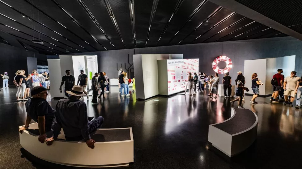 Ein Blick in den Denkraum Technik. Im Hintergrund sind Wände mit Ausstellungstehemen zu sehen. Im Vordergrund sitzen Menschen auf Bänken.