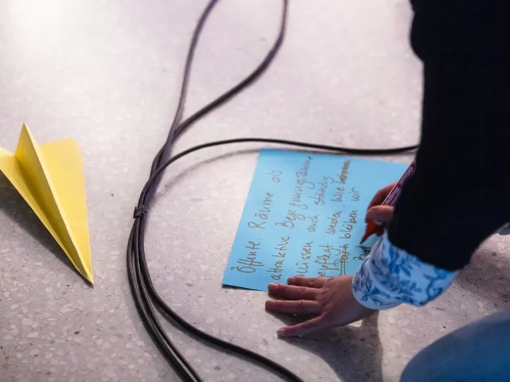 Auf einem Boden liegen bunte Blätter. Eine Hand schreibt etwas auf. Daneben ein Kabel und ein Papierflieger