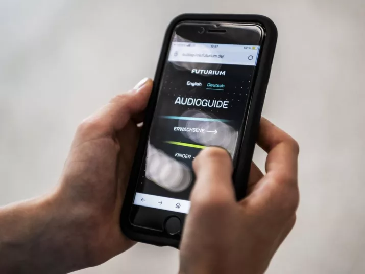 Zwei Hände halten ein Smartphone, auf dem der Audioguide geöffnet ist.